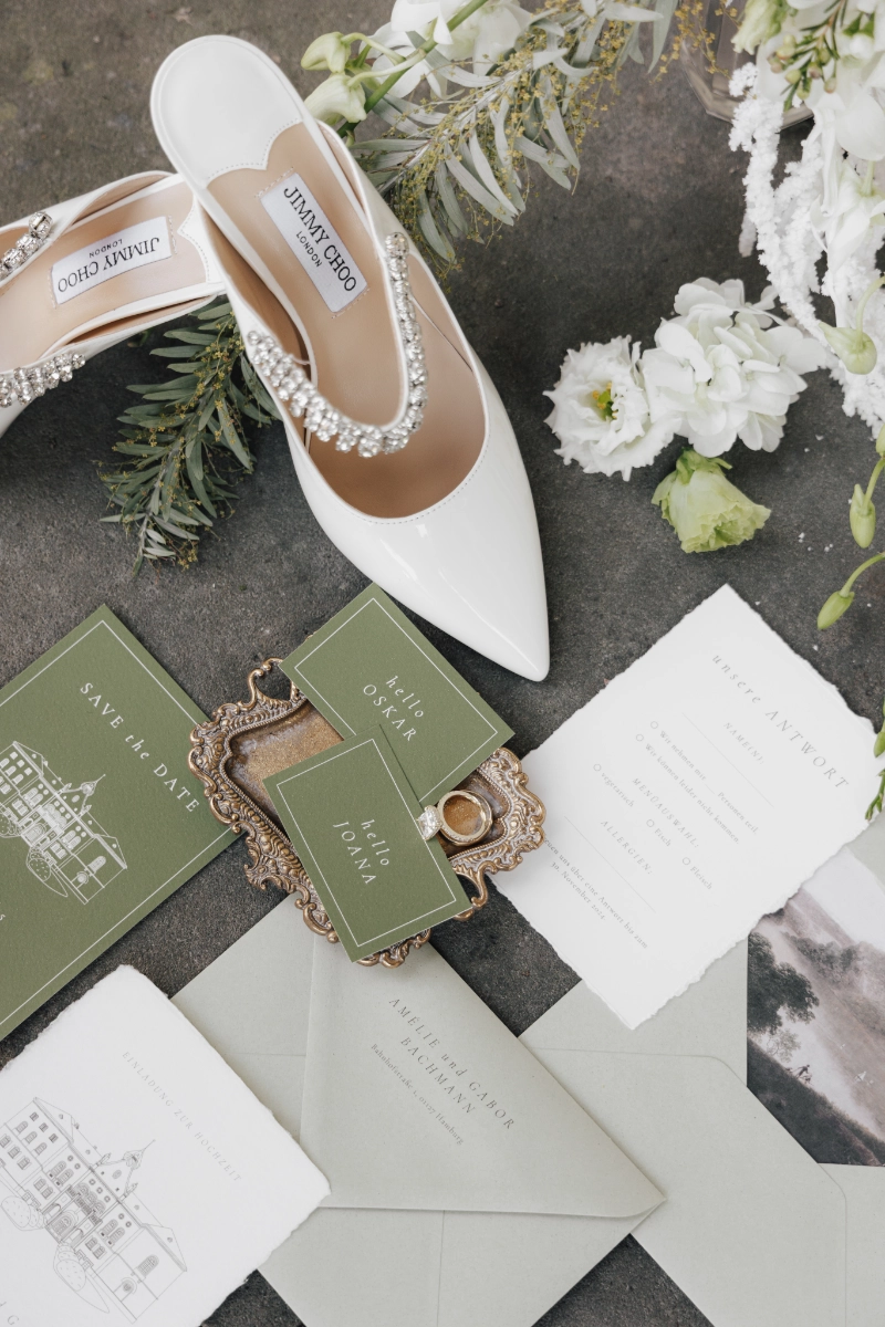 Individuelle Hochzeitseinladung in weiß und grün in einem Flatlay für eine Fine Art Hochzeit aus handgeschöpftem Papier mit einer Hochzeitslocation Zeichnung und weißer Schrift auf Einladung sowie Adressbeschriftung auf Briefumschlag. Brautschuhe von Jimmy Choo